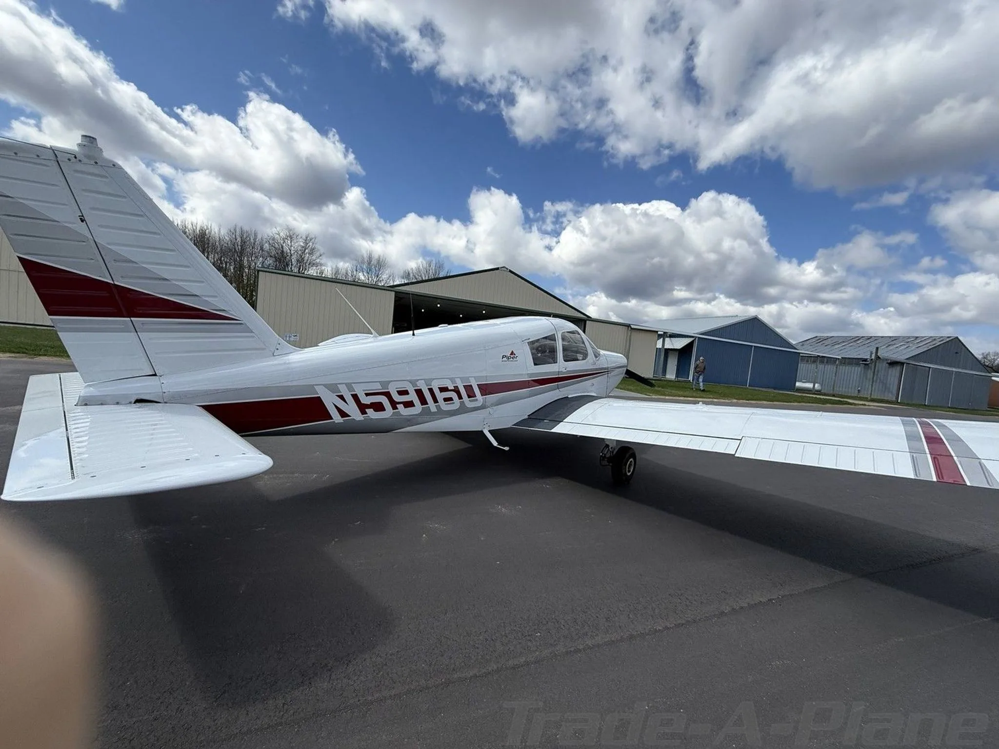 A side view of a white Piper Cherokee aircraft with red and grey stripes on an airfield with hangars in the background.