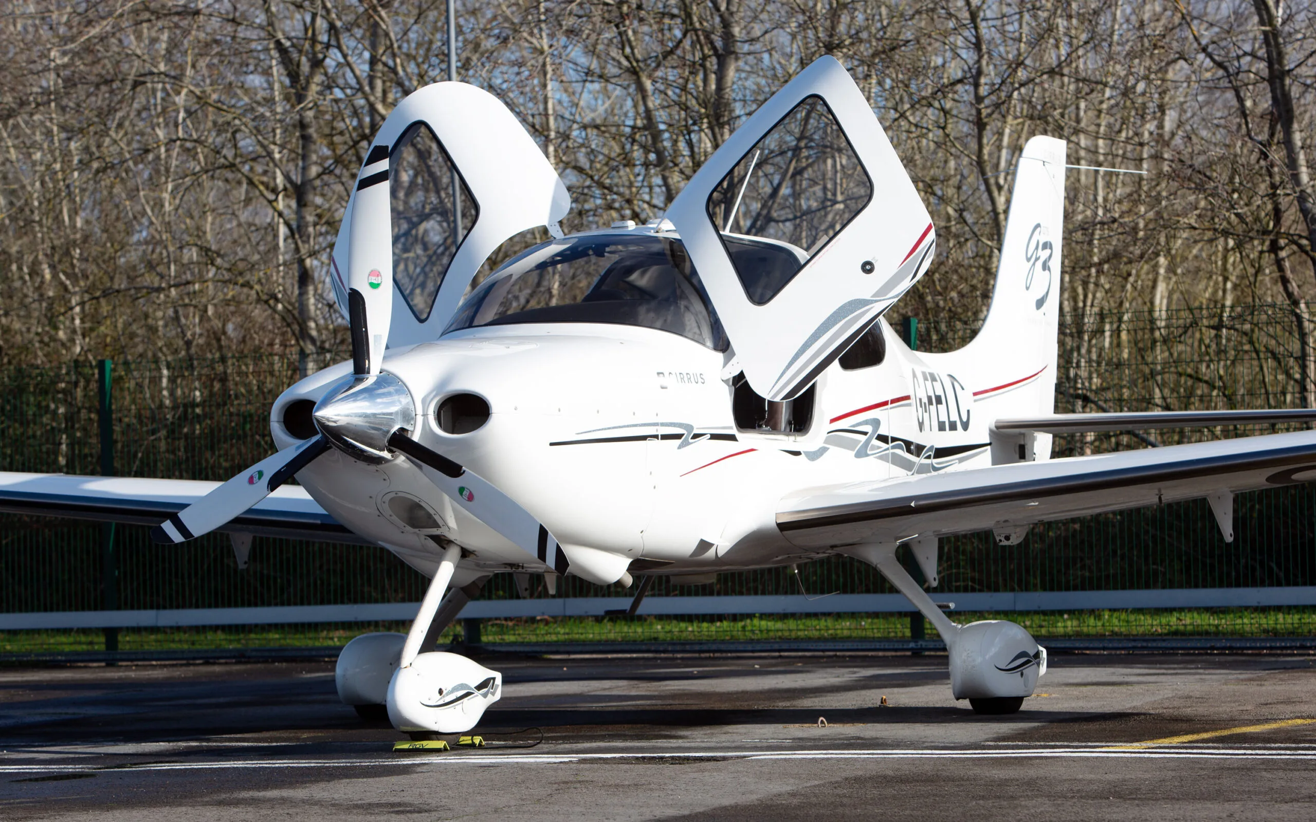 A white Cirrus SR22 G3 aircraft with red and black stripes, registration G-FELC, parked on a tarmac with its gull-wing doors open.