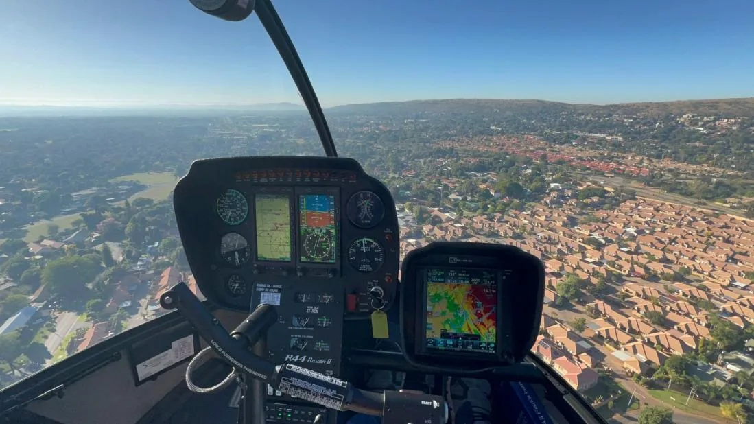 The cockpit of a Robinson R44 helicopter with instruments visible, looking out over a residential area.