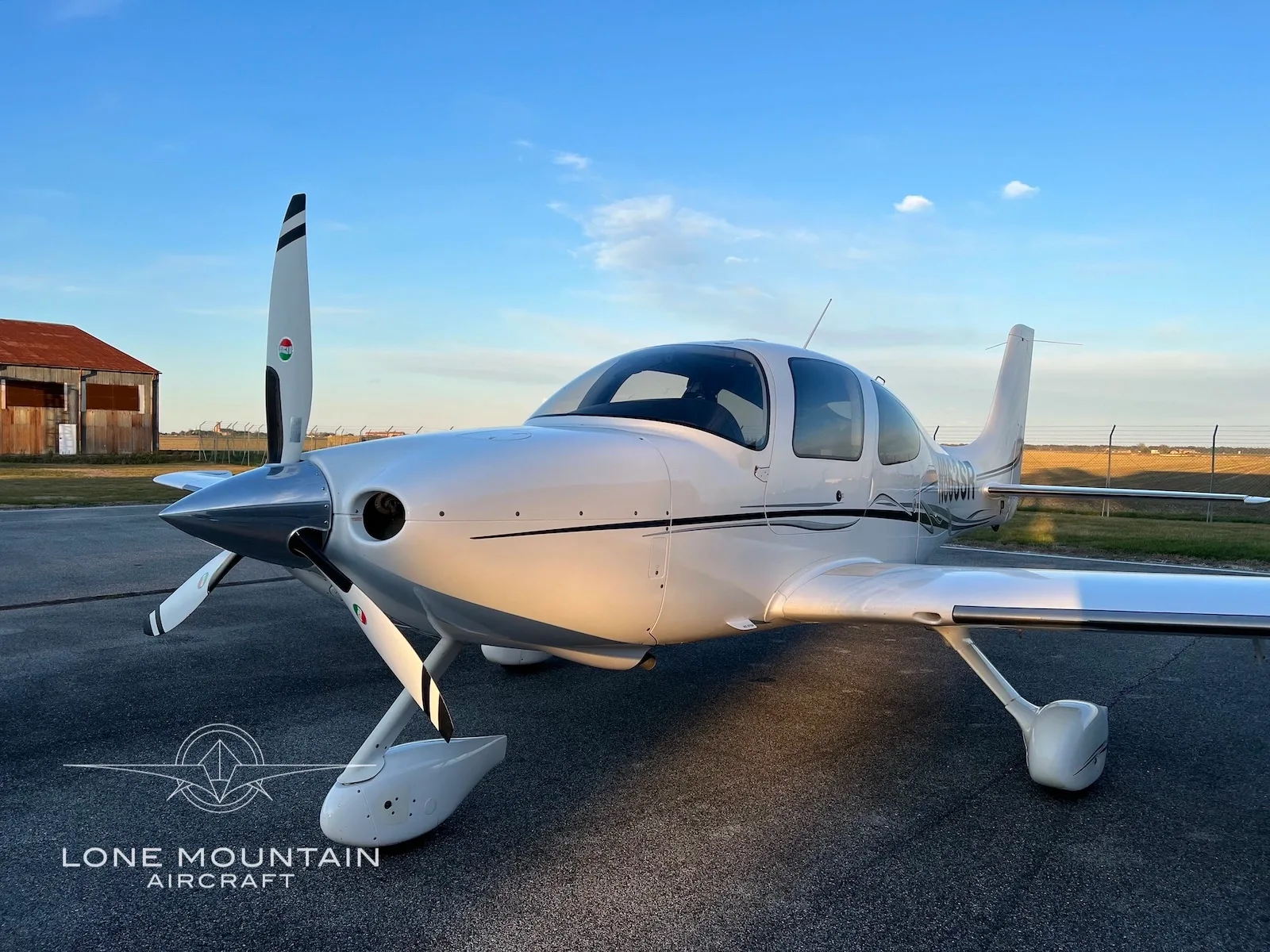 A white Cirrus SR22 aircraft with black and grey stripes is parked on an airfield at sunset, with a rustic building in the background.