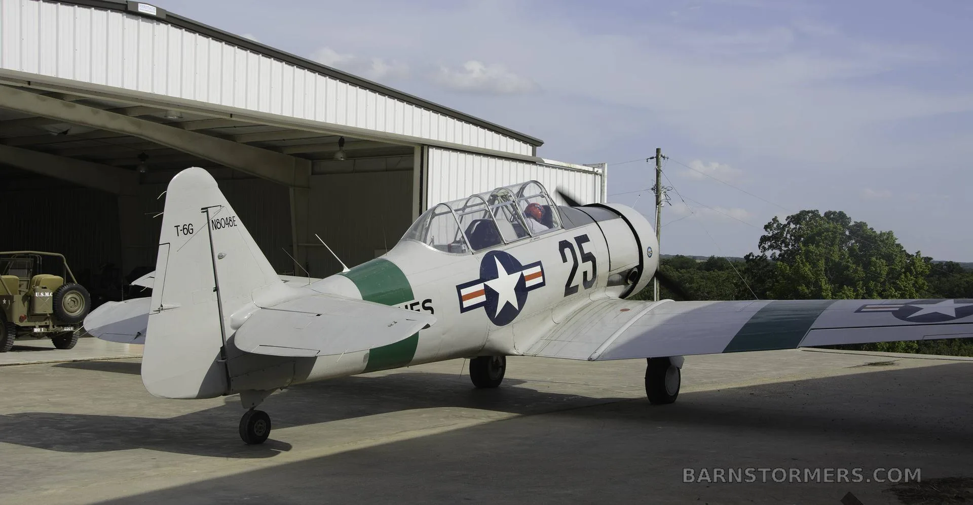 A light gray T-6G Texan aircraft with "25" markings, a US star insignia, and a green stripe, parked on concrete outside a hangar.
