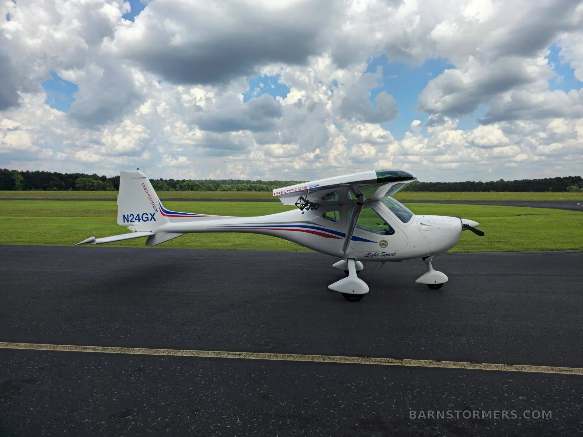 A white Remus GX Light Sport aircraft with red and blue stripes is parked on an asphalt runway, viewed from the rear-left side.