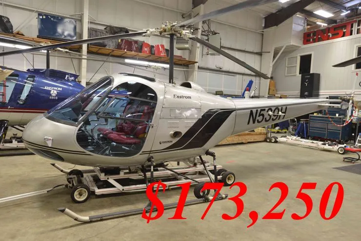 A silver Enstrom helicopter with black stripes and red interior, registration N539H, is parked in a hangar.