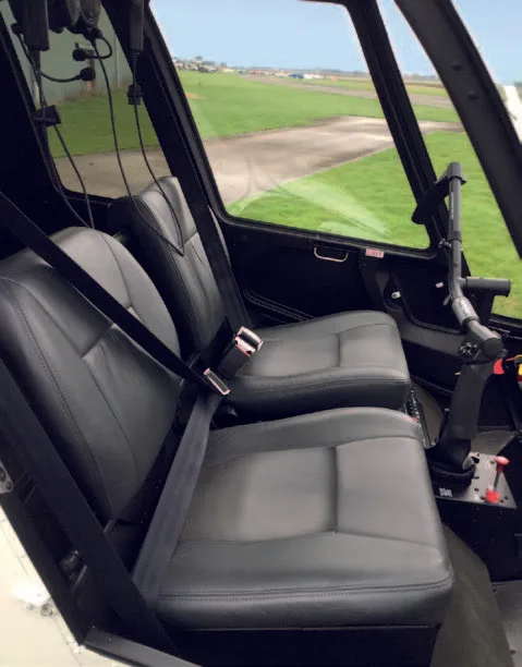 The black leather seats of a helicopter interior, with a view of an airfield through the windows.