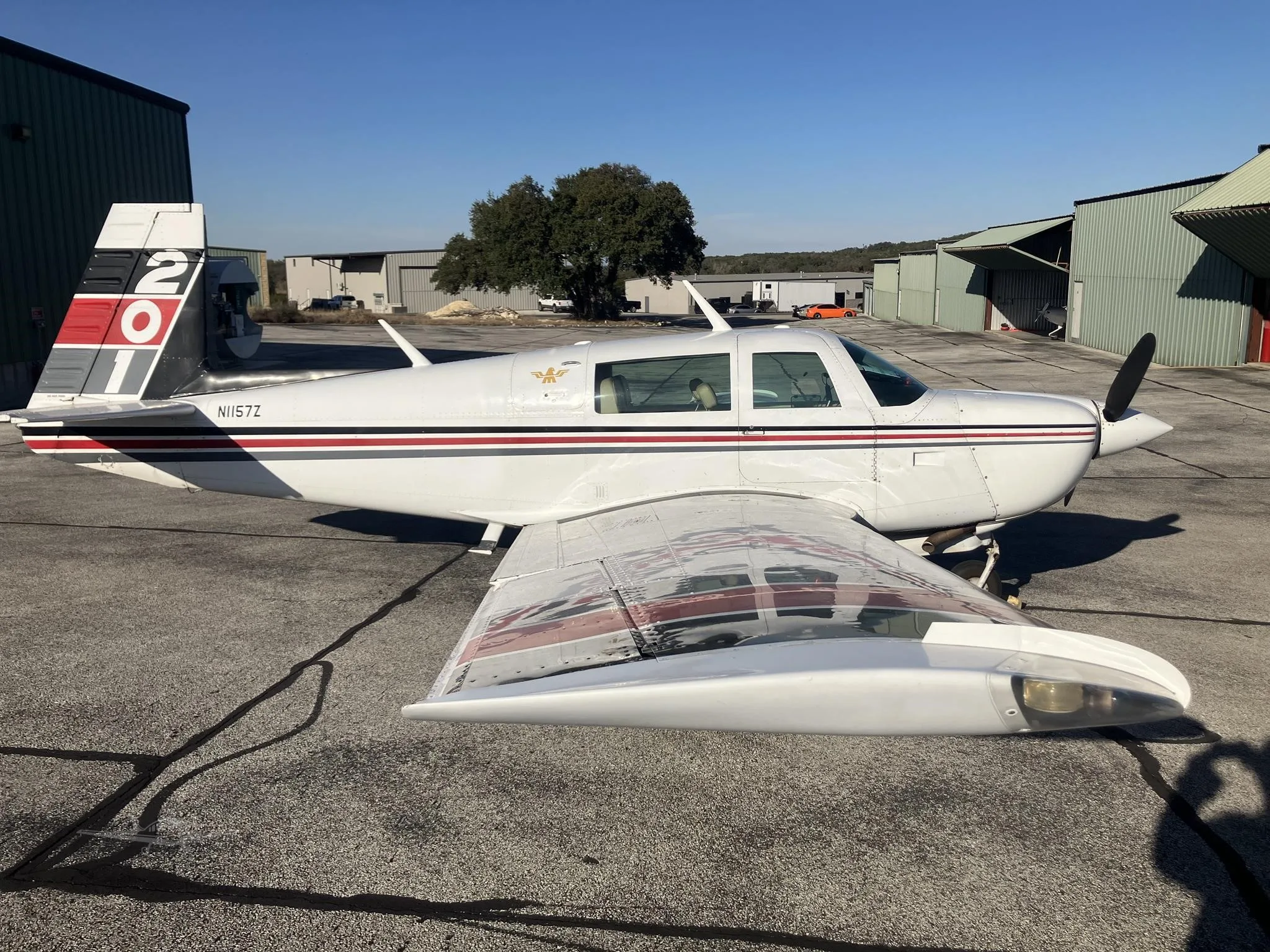 A white single-engine propeller aircraft with red and black stripes is parked on an airfield tarmac with hangars in the background.
