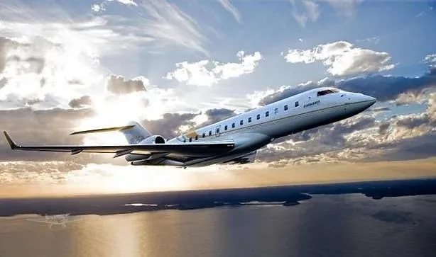 A white Bombardier Global business jet flies over water at sunset, with 'BOMBARDIER' visible on its engine.