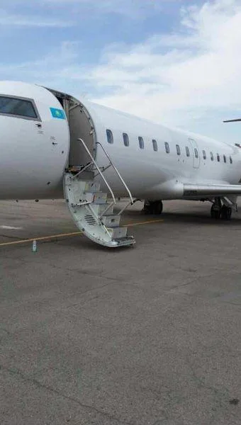 A white regional jet with a blue and yellow flag on its nose, parked on an asphalt tarmac with its main passenger door open and integrated stairs extended.