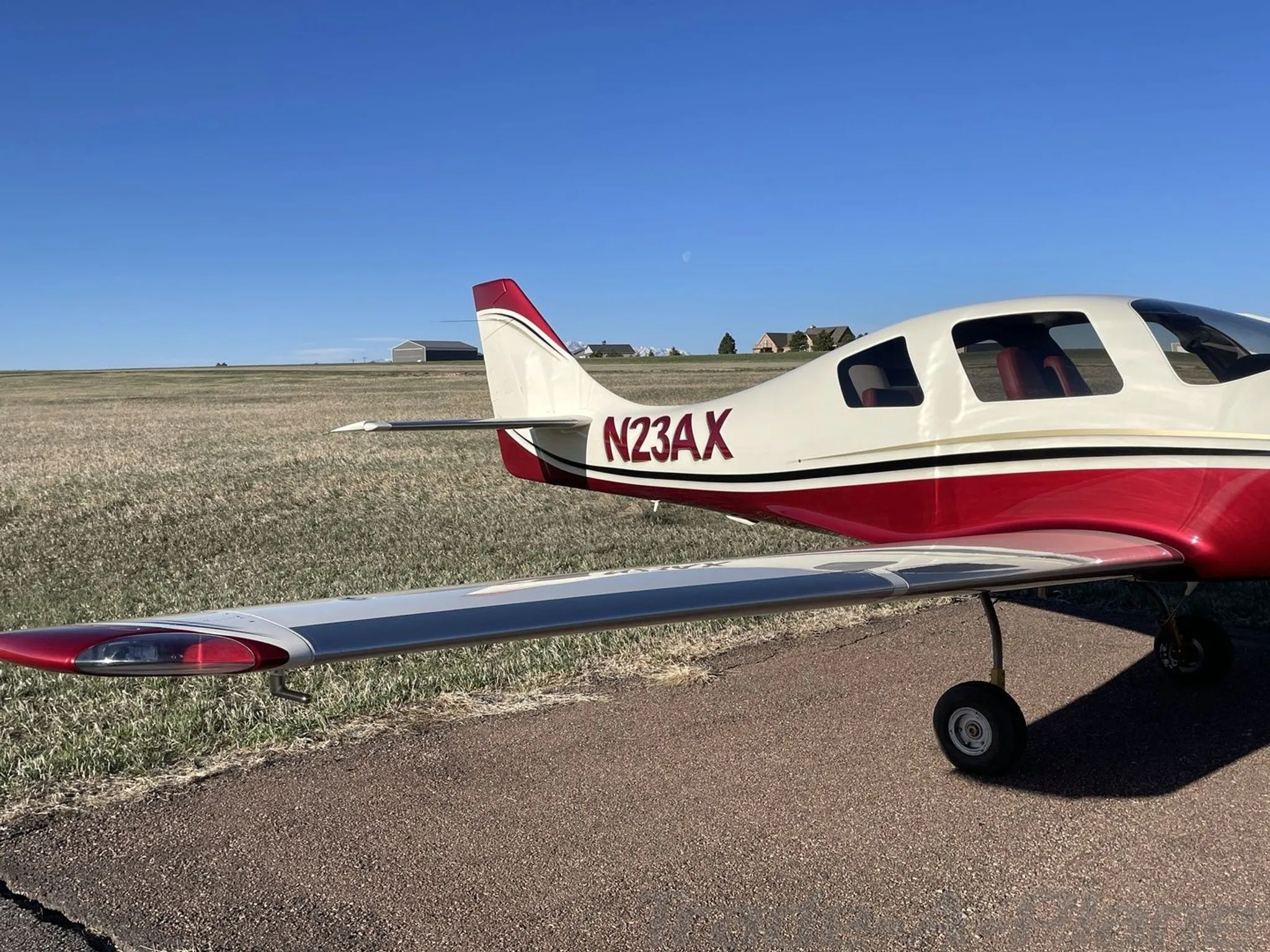 A side view of the white and red aircraft N23AX, showing its tail and wingtip on a paved surface with a field in the background.