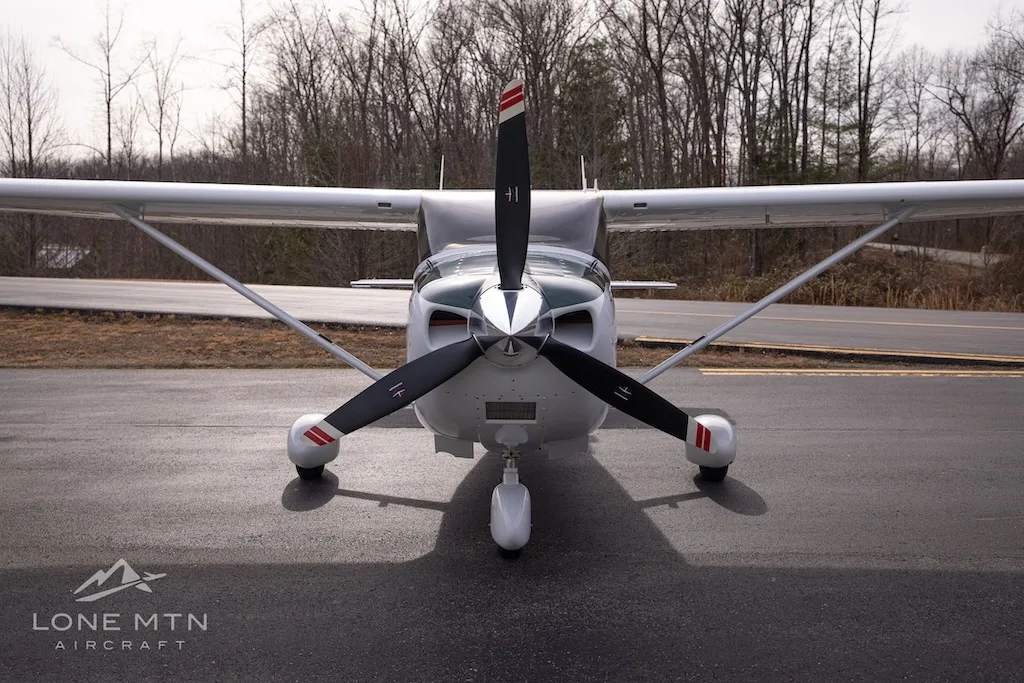 A head-on view of a white and dark green Cessna Skylane N915LS with a three-bladed propeller, parked on an asphalt ramp.