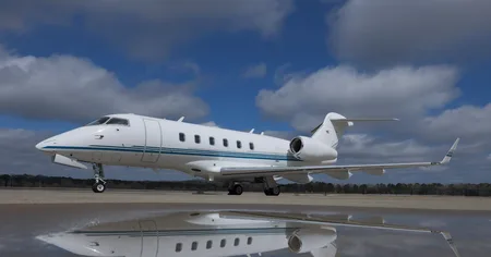 A white business jet with a blue stripe is parked on a wet tarmac, reflecting under a partly cloudy blue sky.