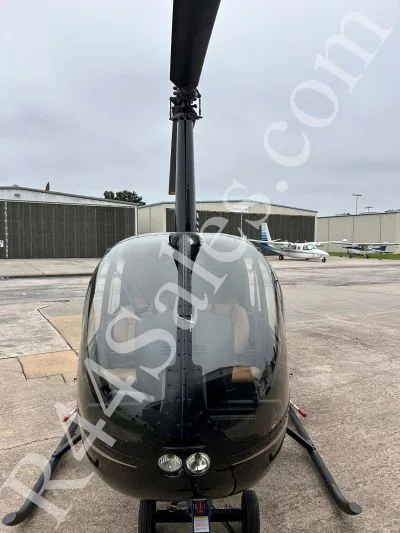 A black Robinson R44 Raven II helicopter with gold pinstripes is parked on a tarmac, viewed from the front.