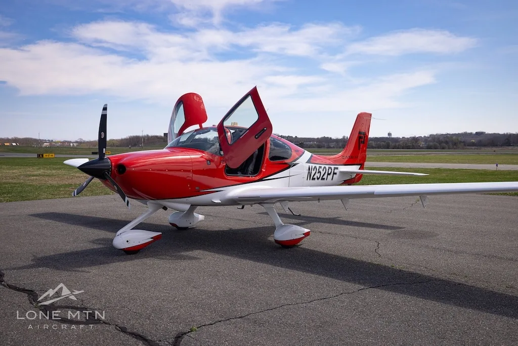 A red and white Cirrus SR20 aircraft with its doors open, parked on an airfield.
