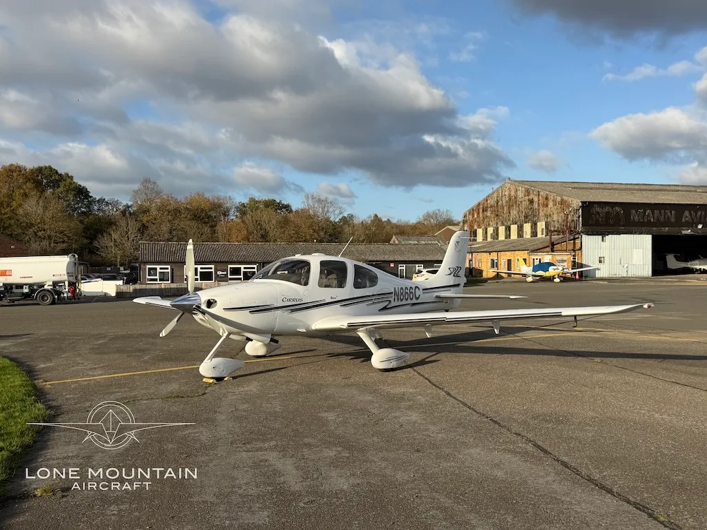 A white Cirrus SR22 aircraft with black and grey stripes, registration N866C, parked on an asphalt apron with hangars in the background.