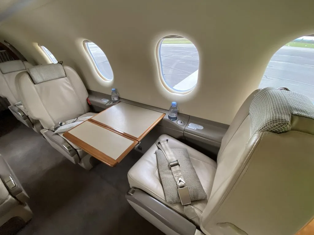 A view inside the aircraft cabin showing two beige leather seats, one with a fold-out table extended between them.
