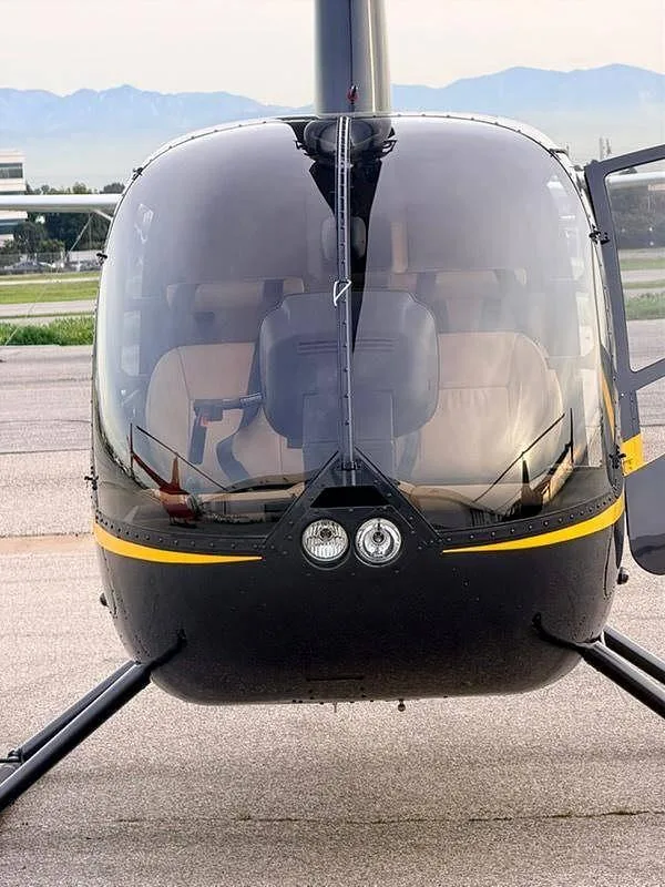 A head-on view of a black Robinson R44 helicopter with a yellow stripe, showing the front windshield and landing lights.
