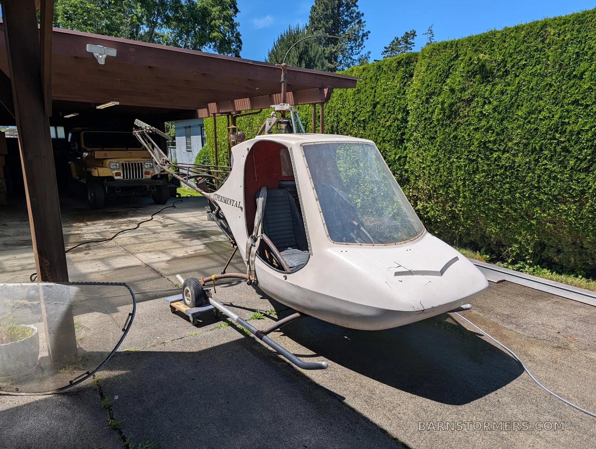 A white experimental Scorpion Too helicopter with an open cockpit, parked on a concrete driveway with a green hedge in the background.