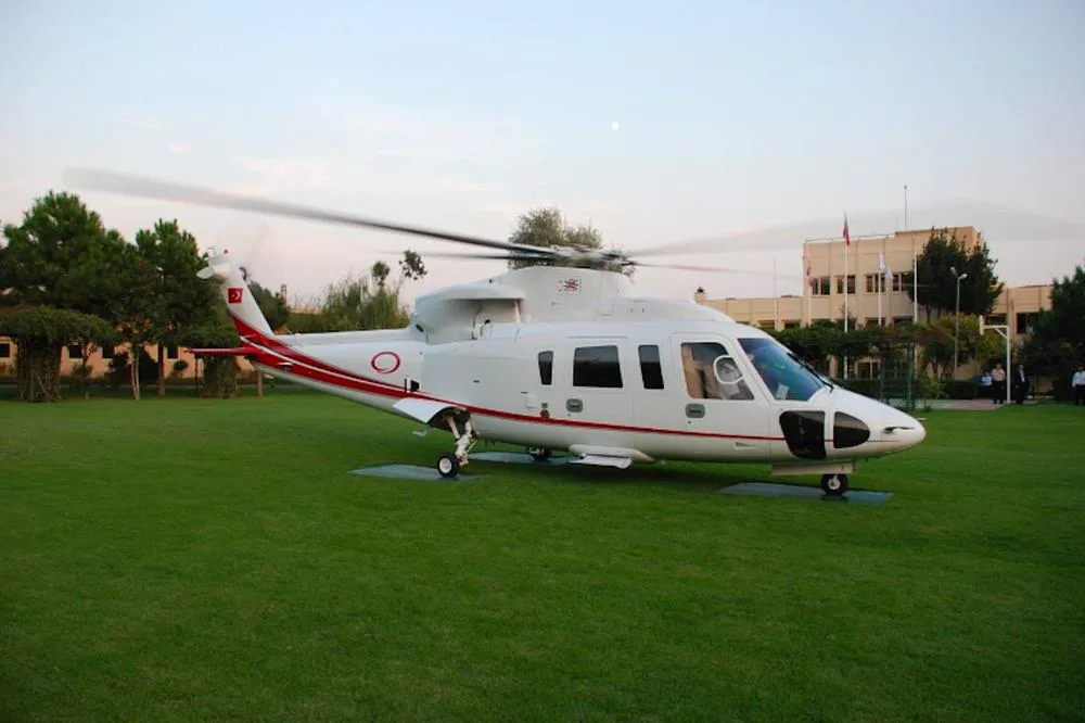 A white helicopter with red stripes and a Turkish flag on the tail, parked on a green lawn in front of a building and trees.