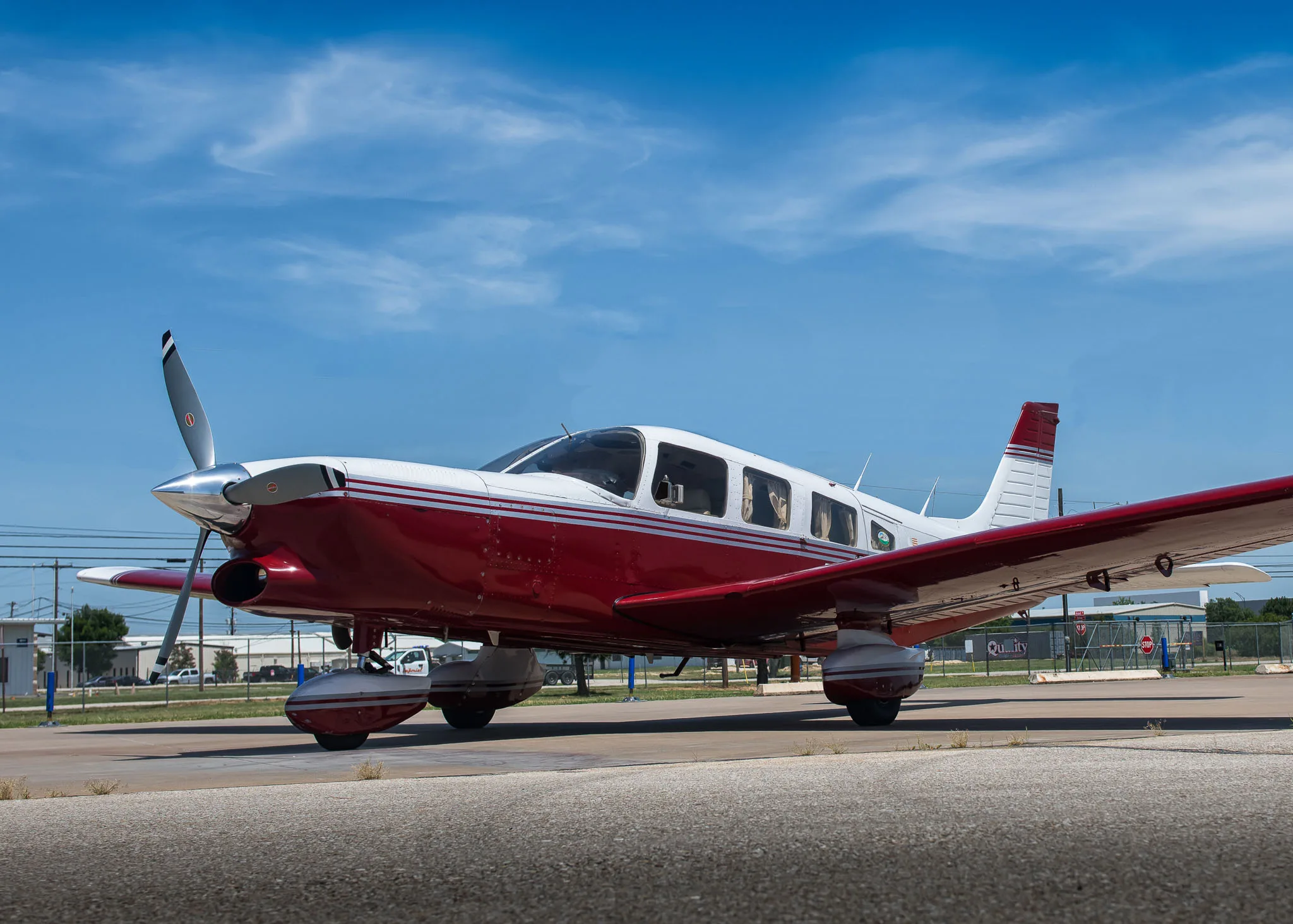 A red and white single-engine aircraft parked on an airport tarmac under a blue sky.