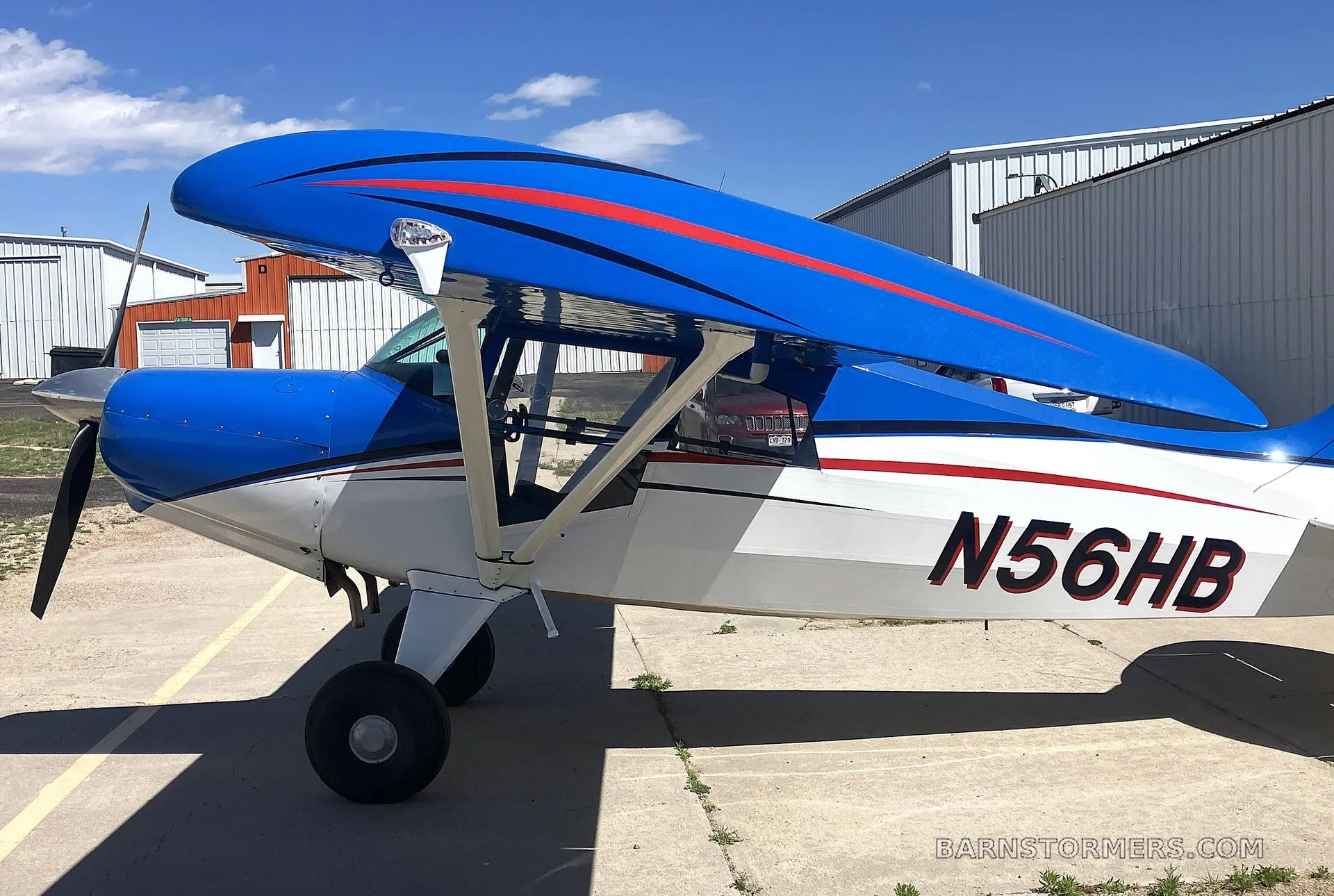 A close-up side view of the front section of a blue and white high-wing aircraft with red stripes, registration N56HB, parked on an apron.