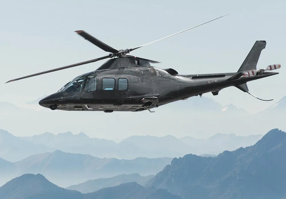 A dark grey helicopter in flight over a range of hazy blue mountains under a clear sky.