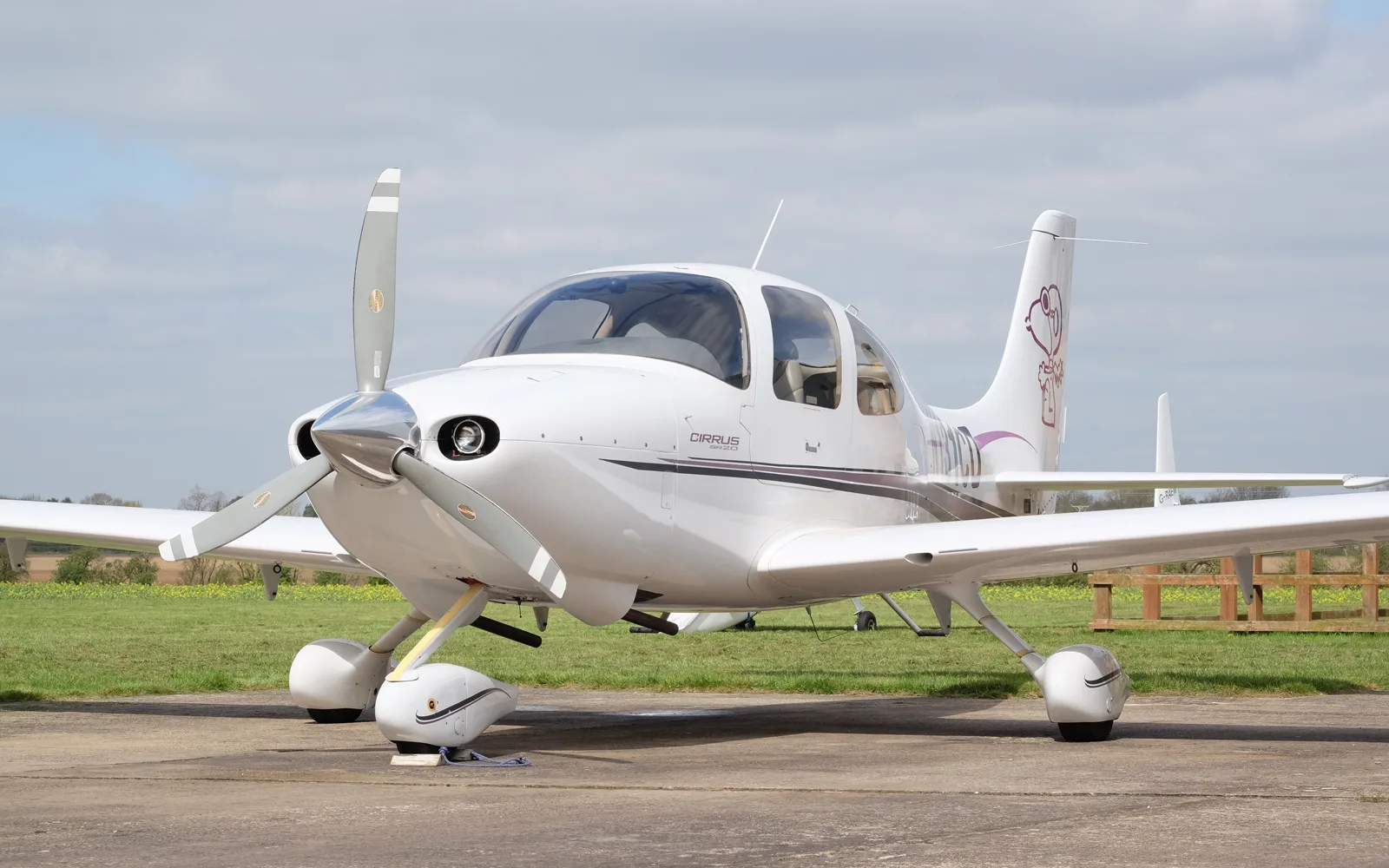 A white Cirrus SR22 aircraft with purple accents and a Snoopy graphic on the tail, parked on a tarmac under a cloudy sky.