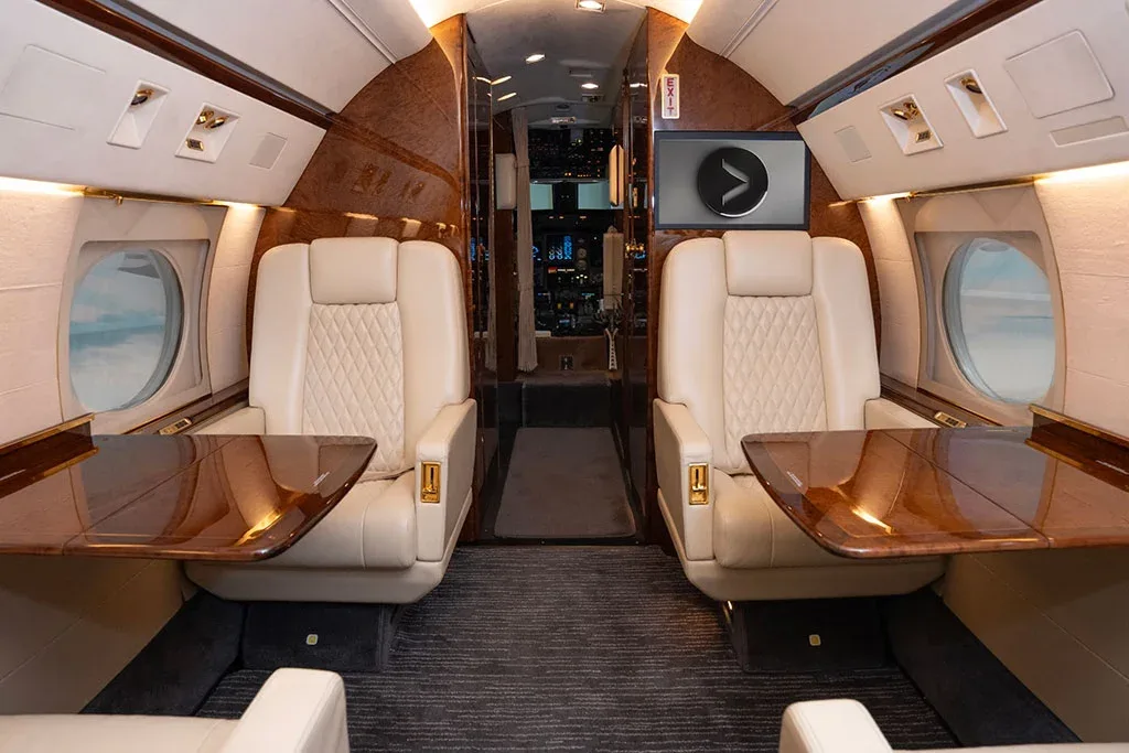 The private jet cabin shows cream leather seats with fold-out polished wood tables.