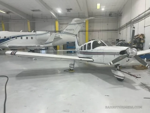 A white single-engine propeller aircraft with black and red stripes parked in a hangar, with a jet aircraft visible in the background.
