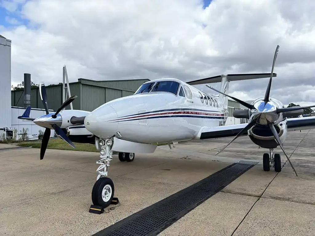 A white twin-turboprop aircraft with red and blue stripes on the fuselage, parked on an asphalt tarmac with hangars in the background.