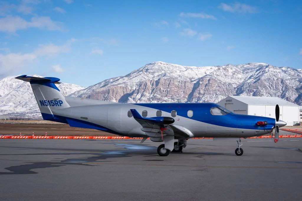 A side view of a blue and silver Pilatus PC-12 aircraft parked on an airfield with snowy mountains and a hangar in the background.