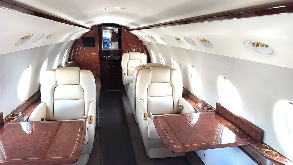 The luxurious interior cabin of a private jet with cream leather seats, polished wood tables, and a galley area at the rear.