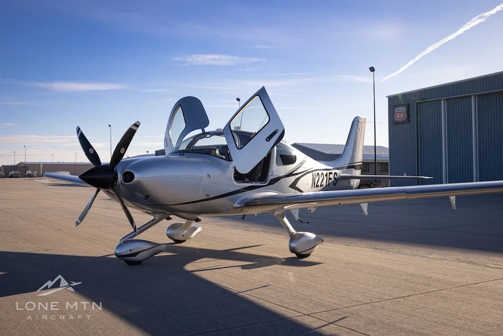A silver Cirrus SR22T aircraft with registration N221FS is parked on an airfield with its doors open.