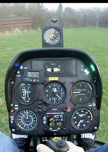The cockpit instrument panel of a Kompress helicopter, showing various gauges and digital displays, with a person's hand on the cyclic control.