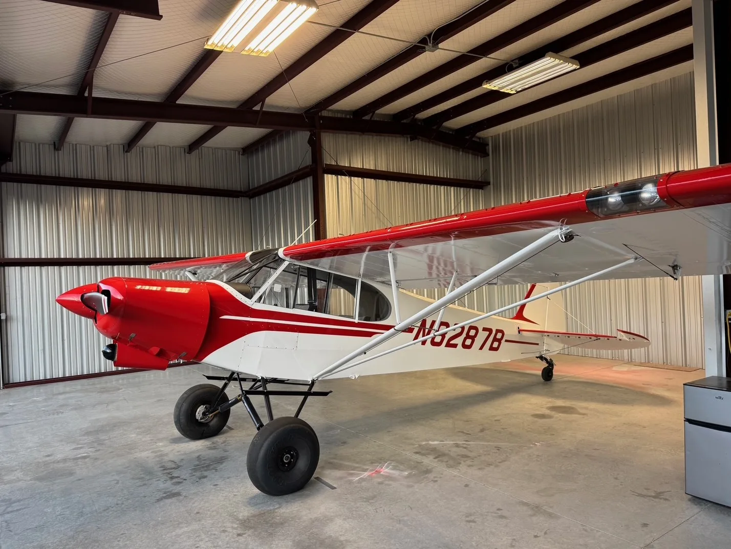 A red and white bush plane, N8287B, with large tires, parked inside a hangar.