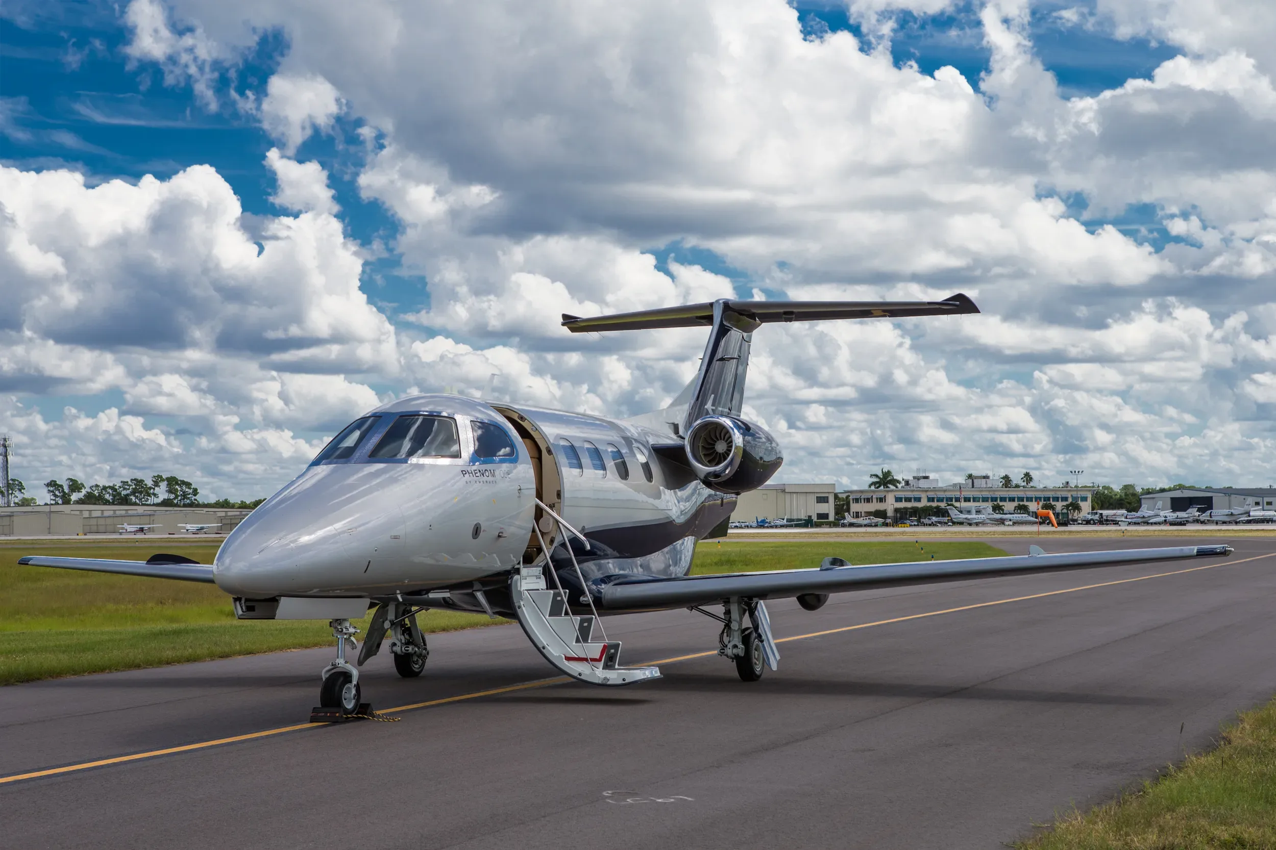 A silver and dark blue Embraer Phenom 100E business jet with its door open and stairs extended, parked on an airport tarmac under a cloudy sky.