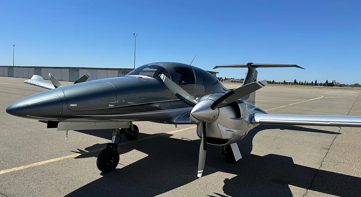A front-quarter view of a grey and silver Diamond DA62 aircraft on an airfield under a clear blue sky.