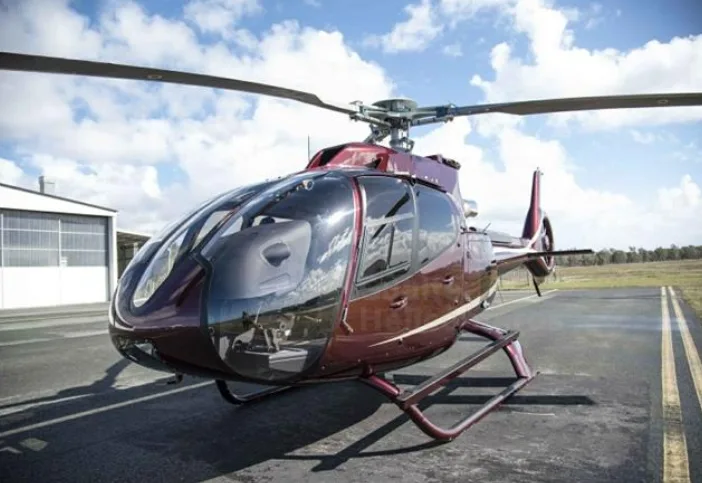 A burgundy helicopter with white stripes parked on an airfield under a partly cloudy sky.