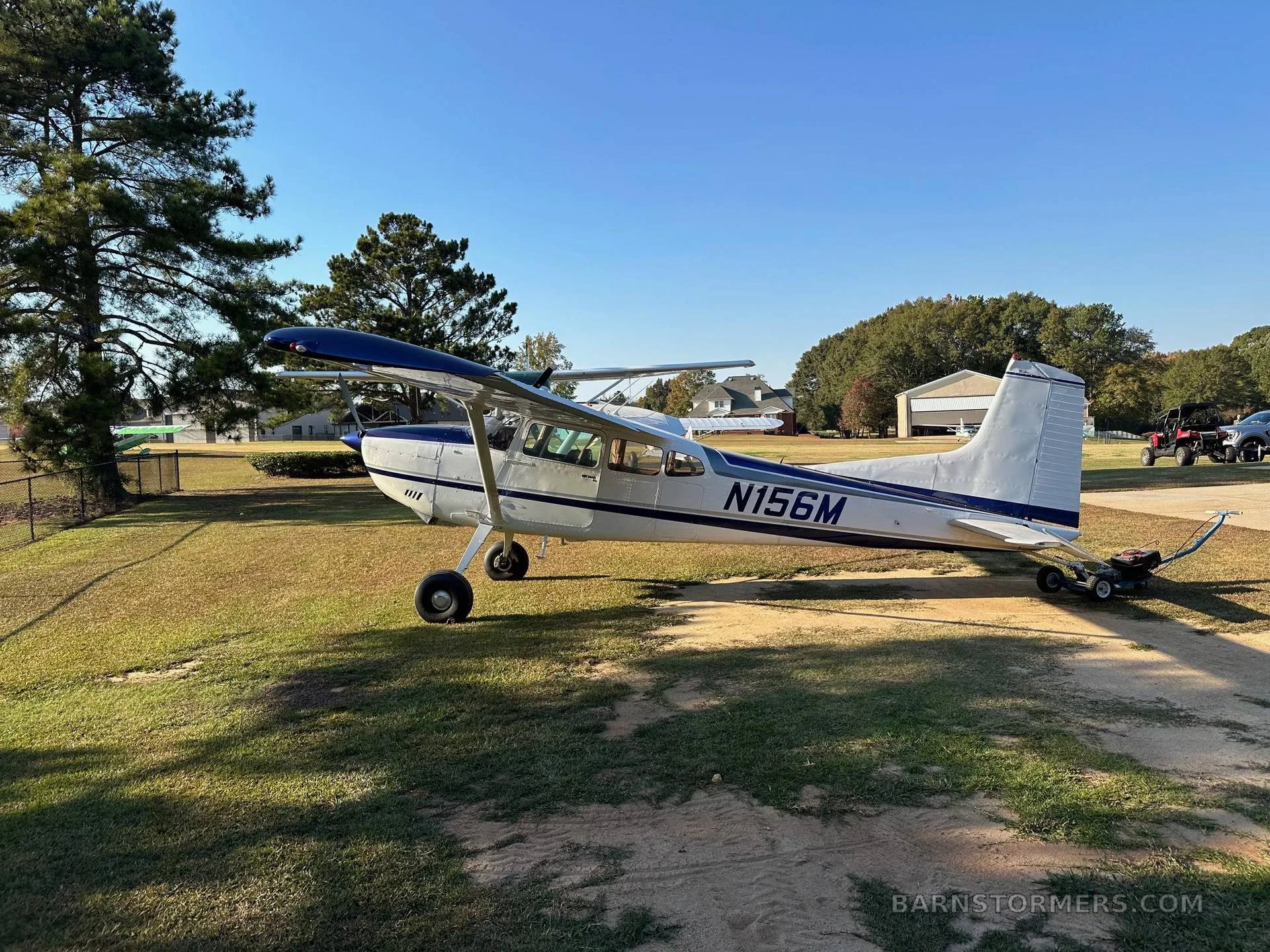 A white and blue Cessna 180 aircraft parked on a sunny grassy field with a building and trees in the background.