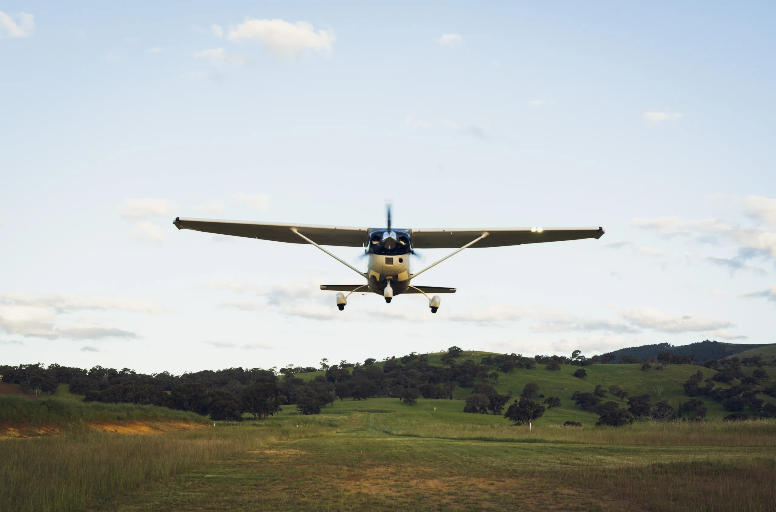 A white single-engine high-wing aircraft with blue accents flying low over a grassy airstrip with rolling hills in the background.