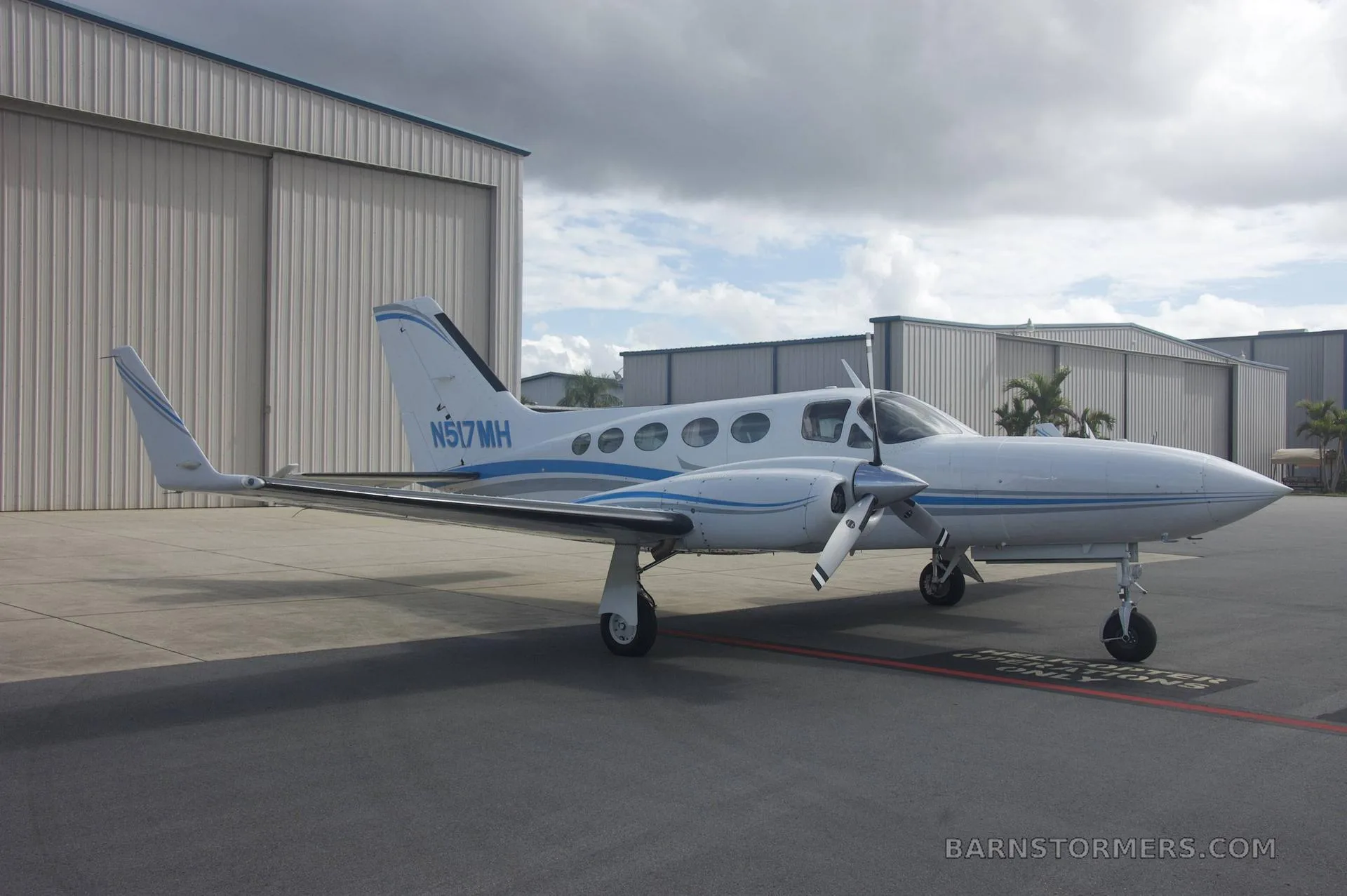 A white twin-engine propeller aircraft with blue and grey stripes (N517MH) is parked on a tarmac next to hangars under a cloudy sky.