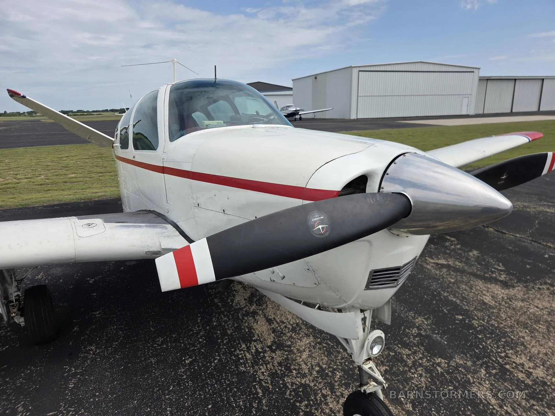 A close-up view of the nose and propeller of a white and red Beechcraft Bonanza aircraft, showing the polished spinner and black propeller blades.