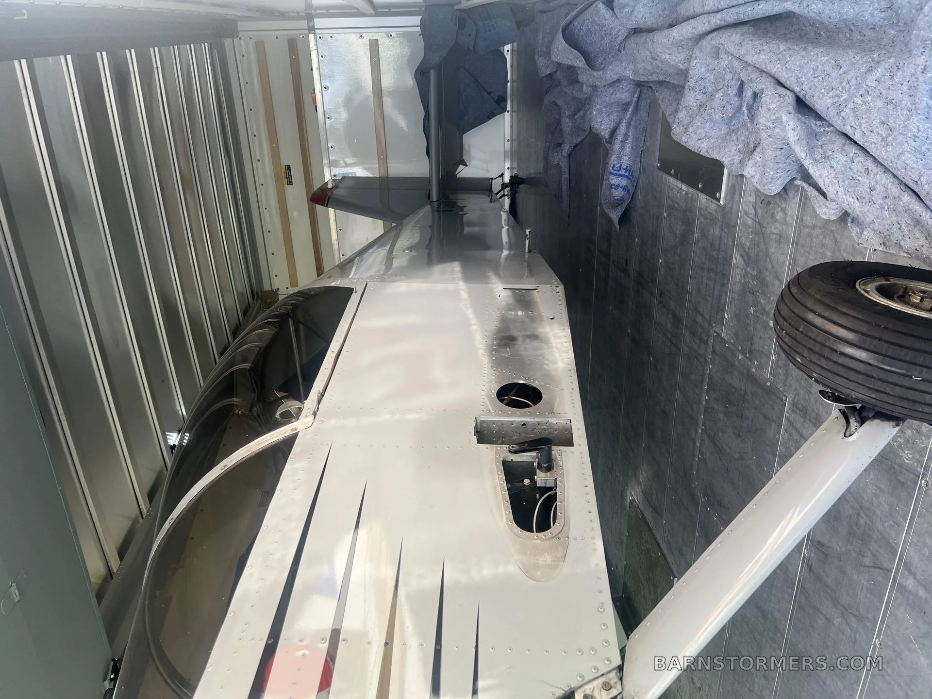 A silver aircraft fuselage with black stripes and a landing gear wheel, partially disassembled inside a trailer.