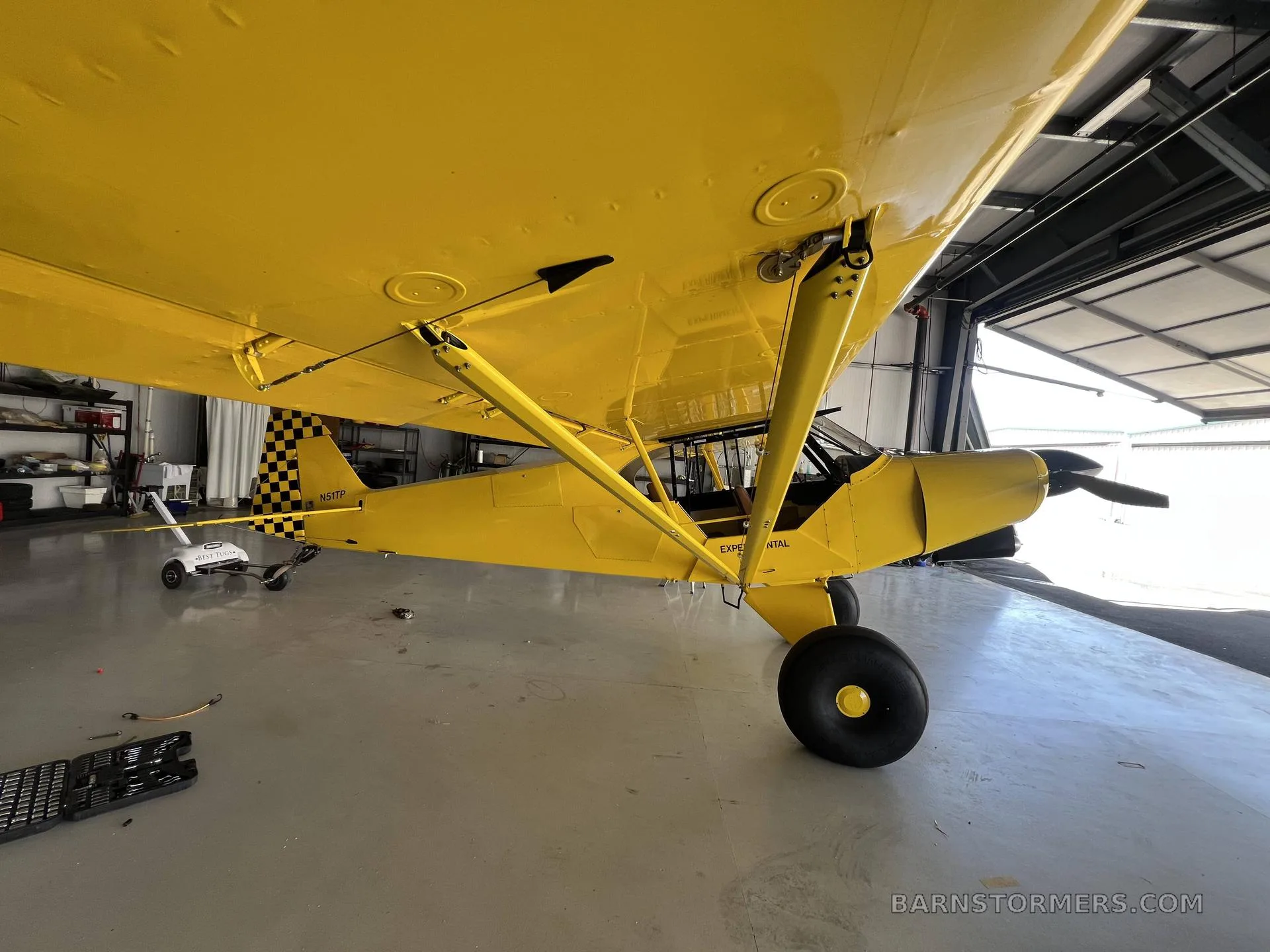 A bright yellow taildragger aircraft, viewed from below the wing, partially inside a hangar.