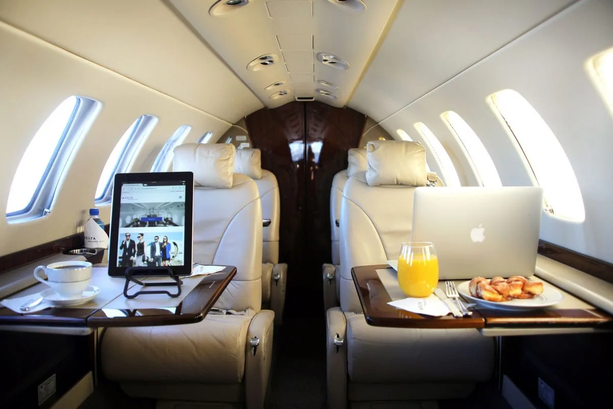 The interior cabin of a private jet, featuring beige leather seats, fold-out tables with a laptop, a tablet, and breakfast items.