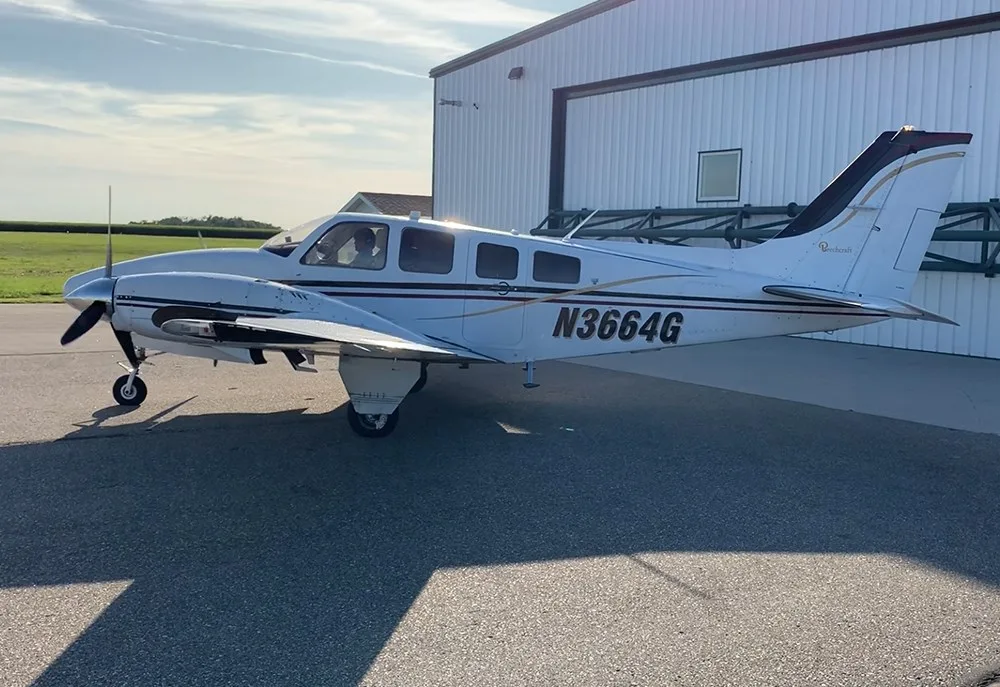 A white Beechcraft Baron N3664G with gold and black stripes is parked on an asphalt ramp next to a white hangar.