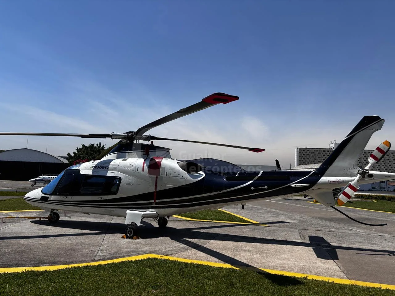 A side view of a white and black Agusta A109 Power helicopter, parked on an airfield under a clear sky.