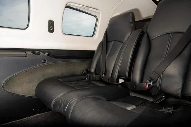 The aircraft cabin features two rows of black leather passenger seats with seatbelts and small windows.