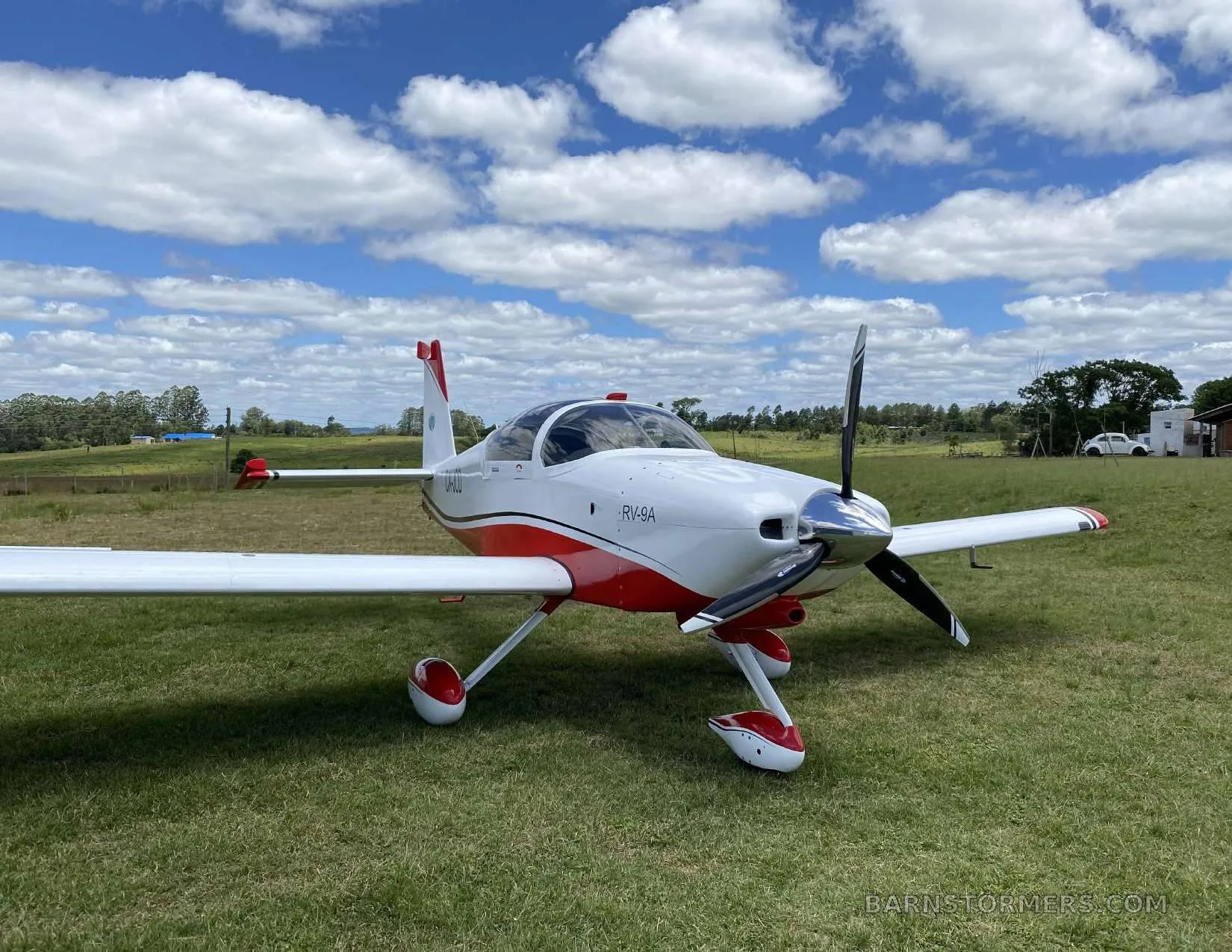 A white and red Van's RV-9A aircraft with a propeller, parked on a grassy field under a blue sky with white clouds.