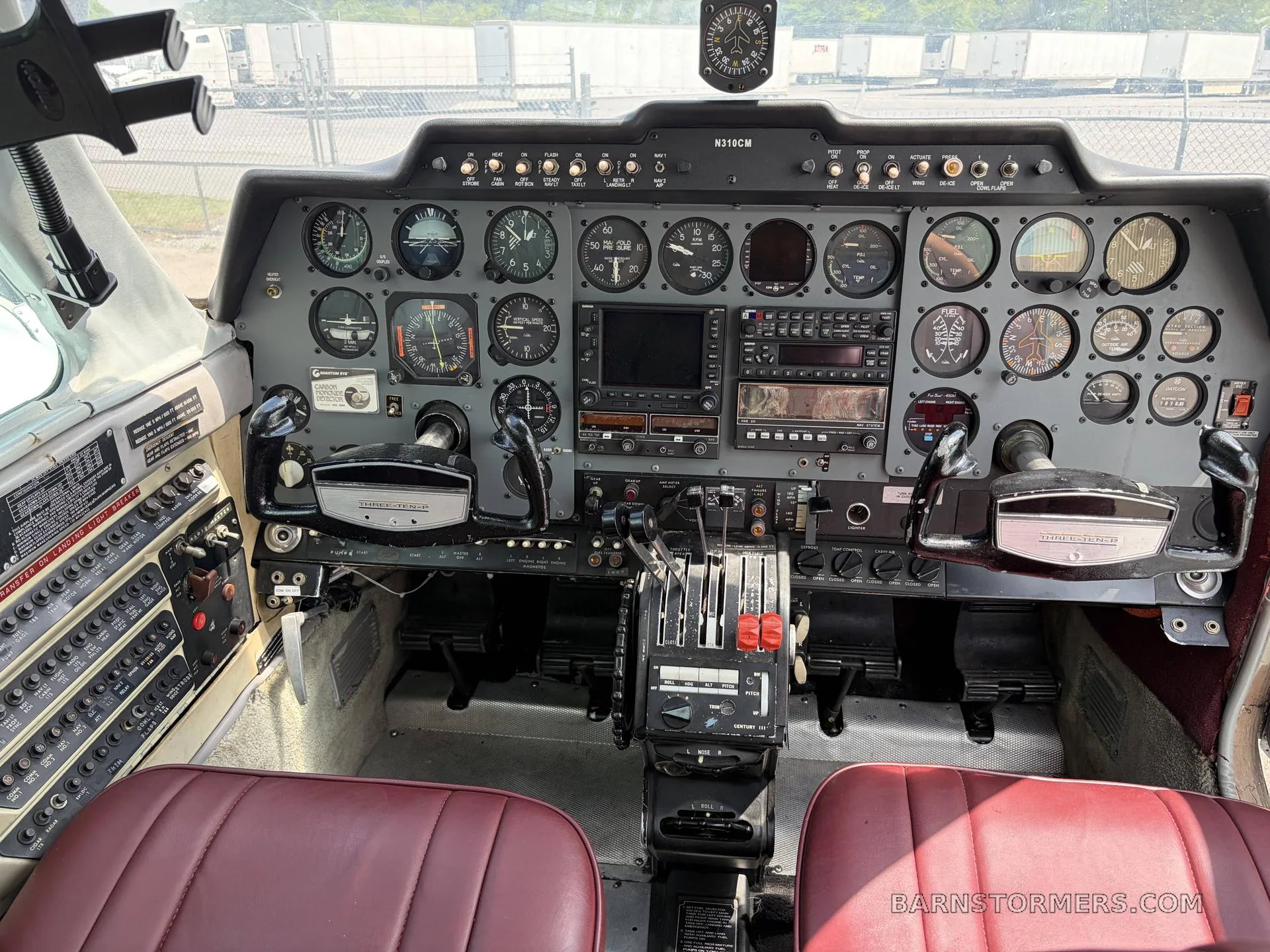 The cockpit of a twin-engine aircraft, showing the instrument panel, yokes, and red leather seats.