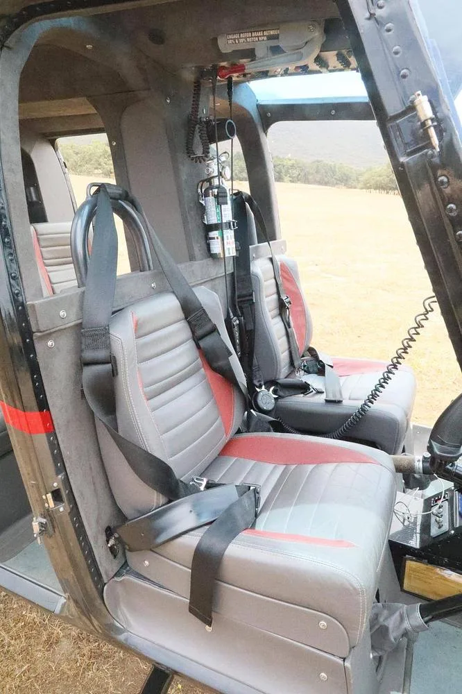 The front two grey and red seats of a helicopter cabin are visible, with seatbelts and a view towards the rear.
