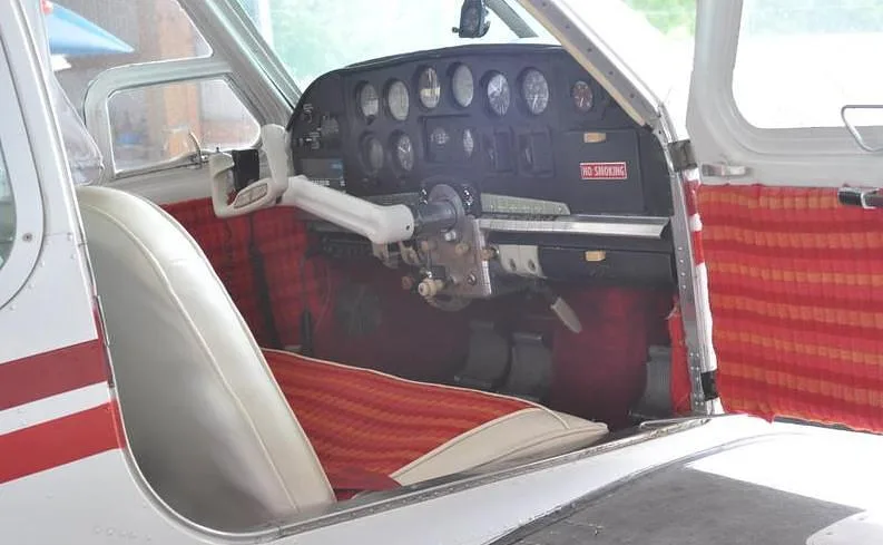 The cockpit of a small aircraft features a white yoke, a full instrument panel, and red patterned interior trim.